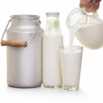 Milk being poured from a pitcher into a glass, with a bottle and metal container on a white background.