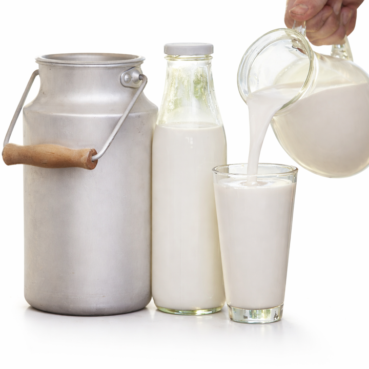 Milk being poured from a pitcher into a glass, with a bottle and metal container on a white background.